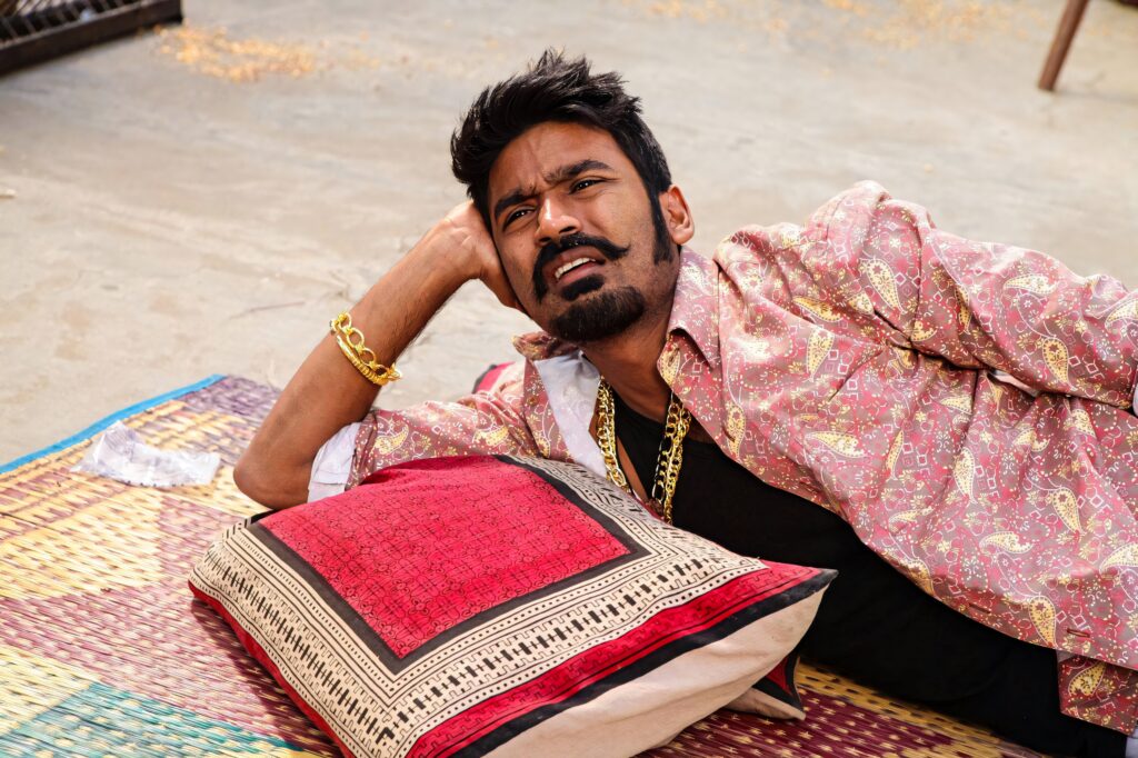 Dhanush lounging on traditional mat wearing patterned shirt and gold chain gritty action vibe wallpaper