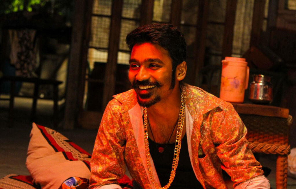 Dhanush as Maari smiling playfully in colorful shirt and gold chain vibrant wallpaper background