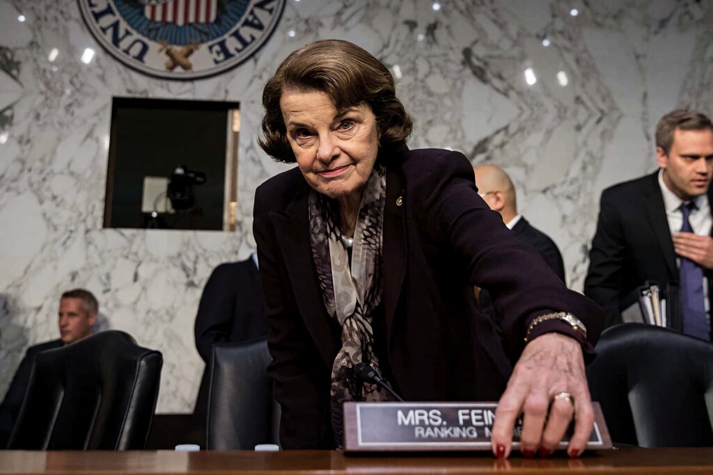 Dianne Feinstein in dark suit with scarf reaching forward in formal political setting wallpaper