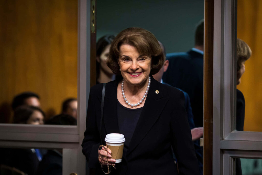 Dianne Feinstein smiling in dark suit pearl necklace holding coffee in warm professional setting wallpaper