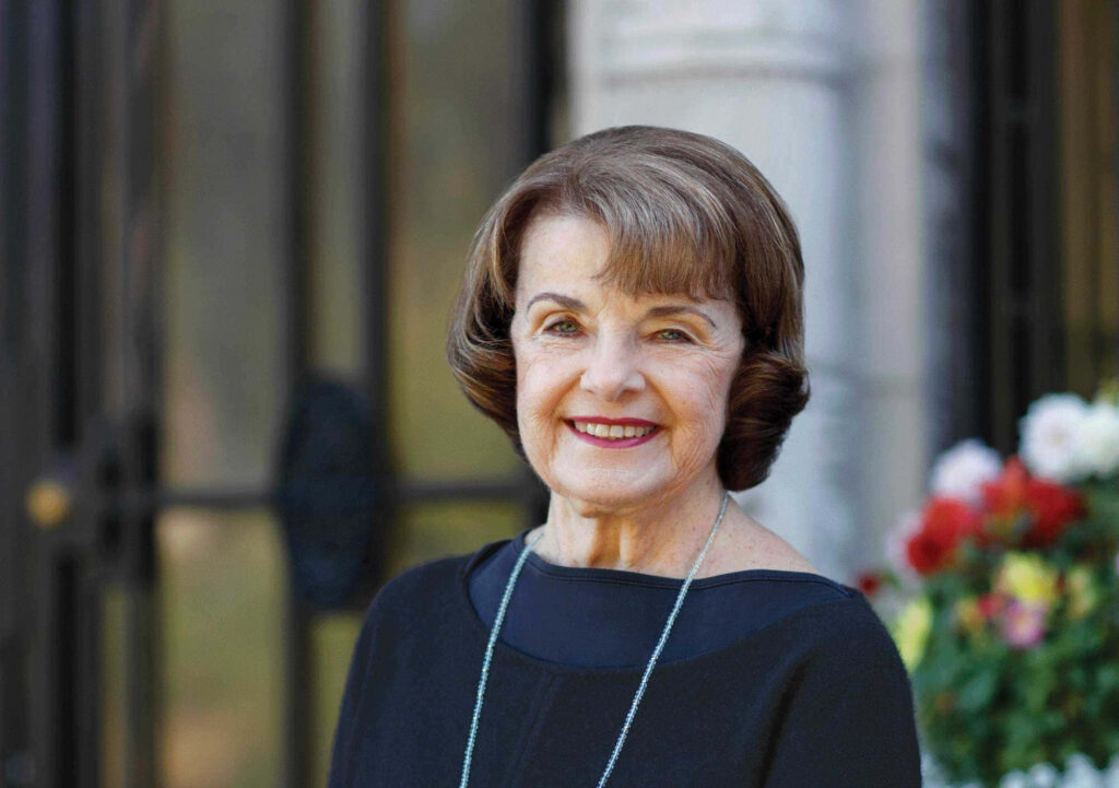 Dianne Feinstein smiling outdoors with iron gate and vibrant flowers wallpaper background
