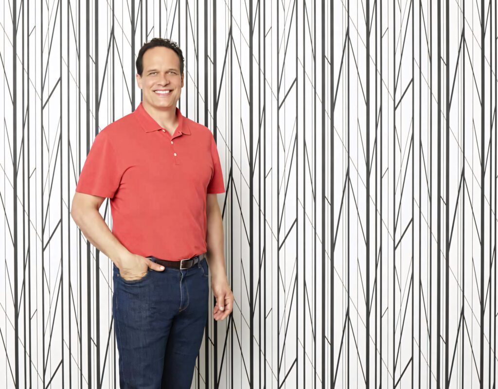 Diedrich Bader in red polo and jeans posing against modern abstract vertical line wallpaper