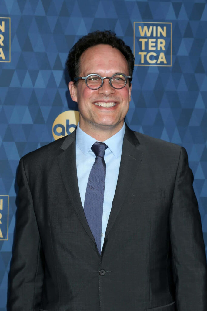 Diedrich Bader smiling in dark suit and light blue tie against blue winter event background Wallpaper