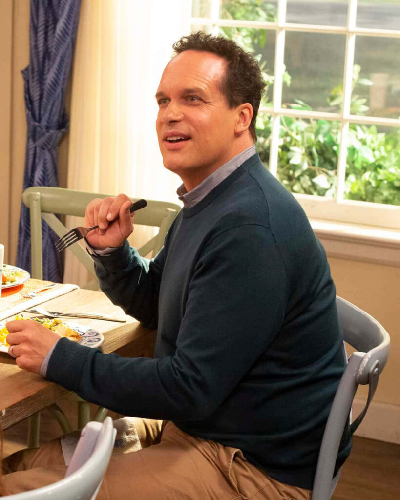 Diedrich Bader sitting at sunlit kitchen table wearing dark sweater and khaki pants with greenery background Wallpaper