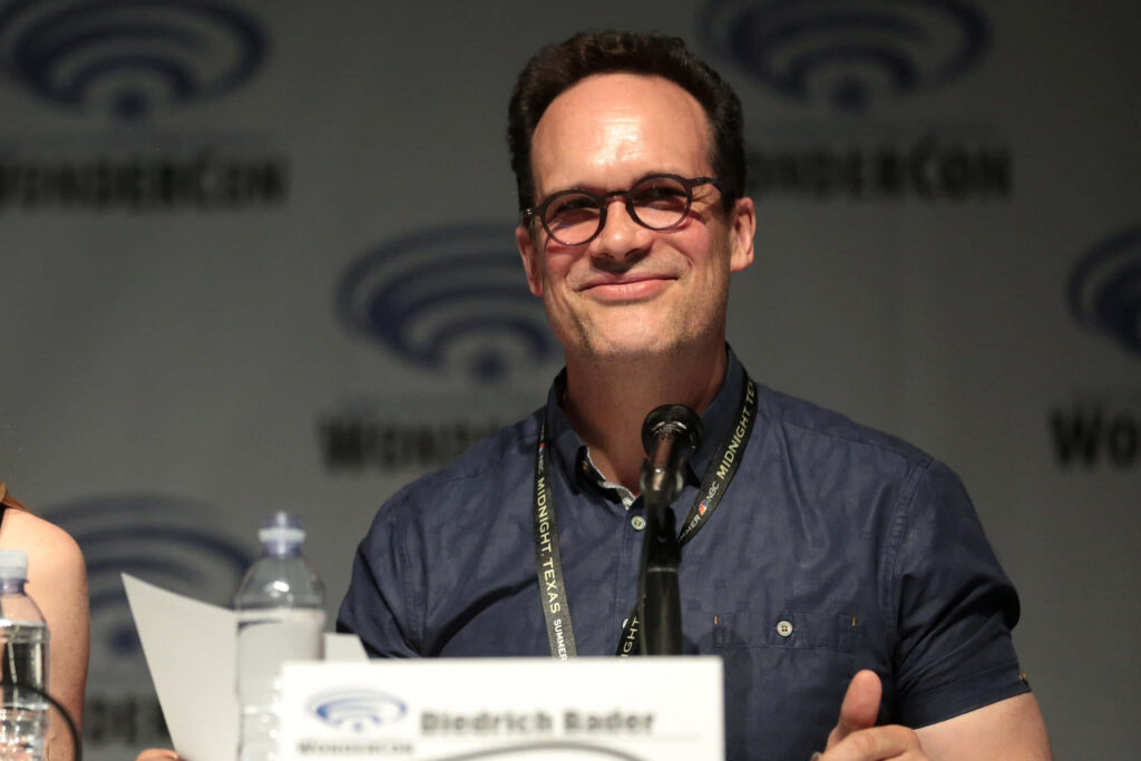 Diedrich Bader smiling at fan convention panel with microphone and branded backdrop lighting Wallpaper