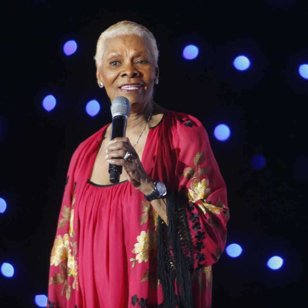 Dionne Warwick on stage in red gold floral robe holding microphone with blue lights background Wallpaper