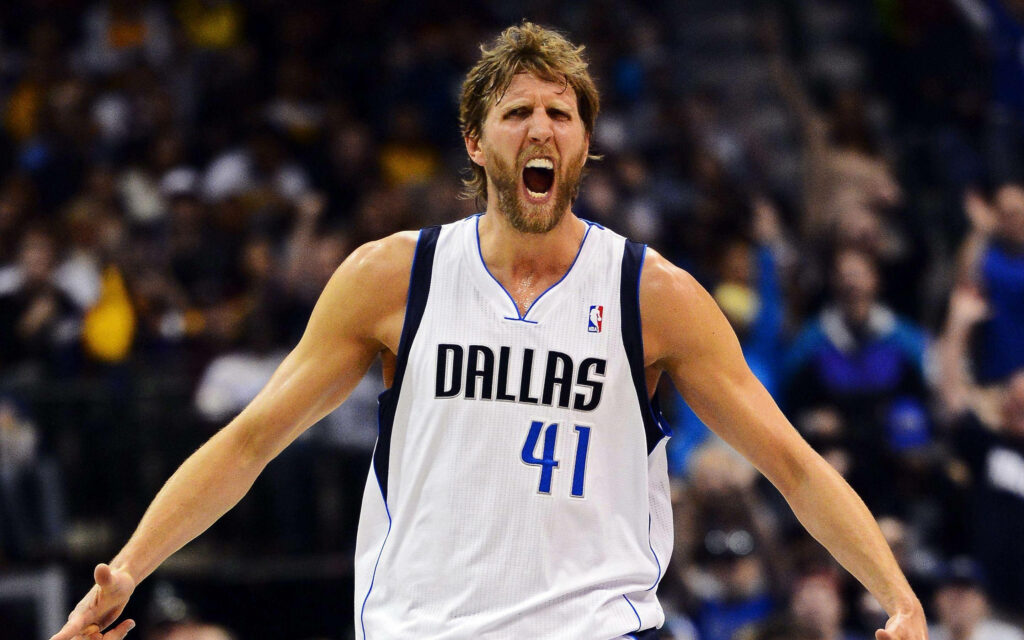 Dirk Nowitzki mid-roar in Dallas Mavericks jersey action shot with intense expression and stadium background Wallpaper