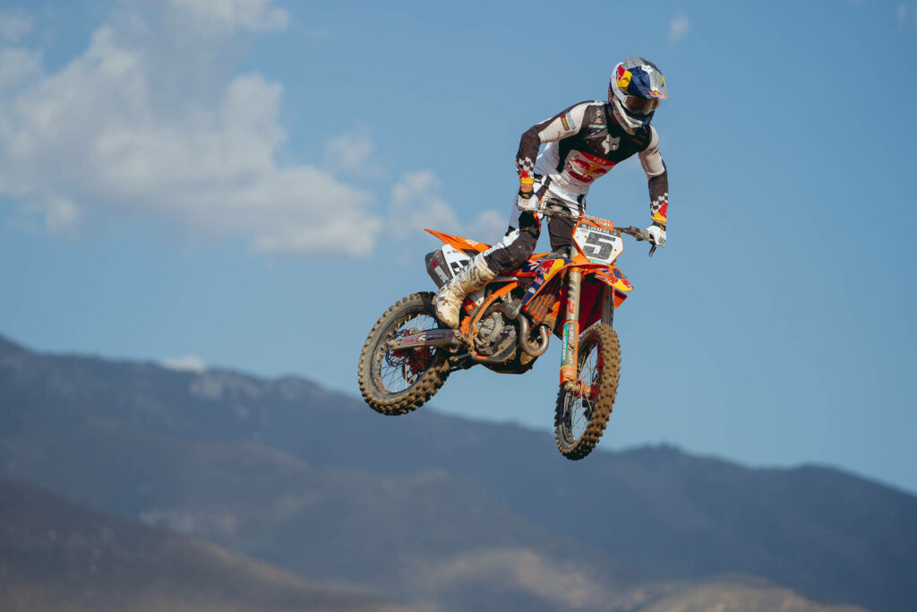 Dirt bike rider soaring over rugged terrain with mountains blue sky vibrant orange motocross action Wallpaper