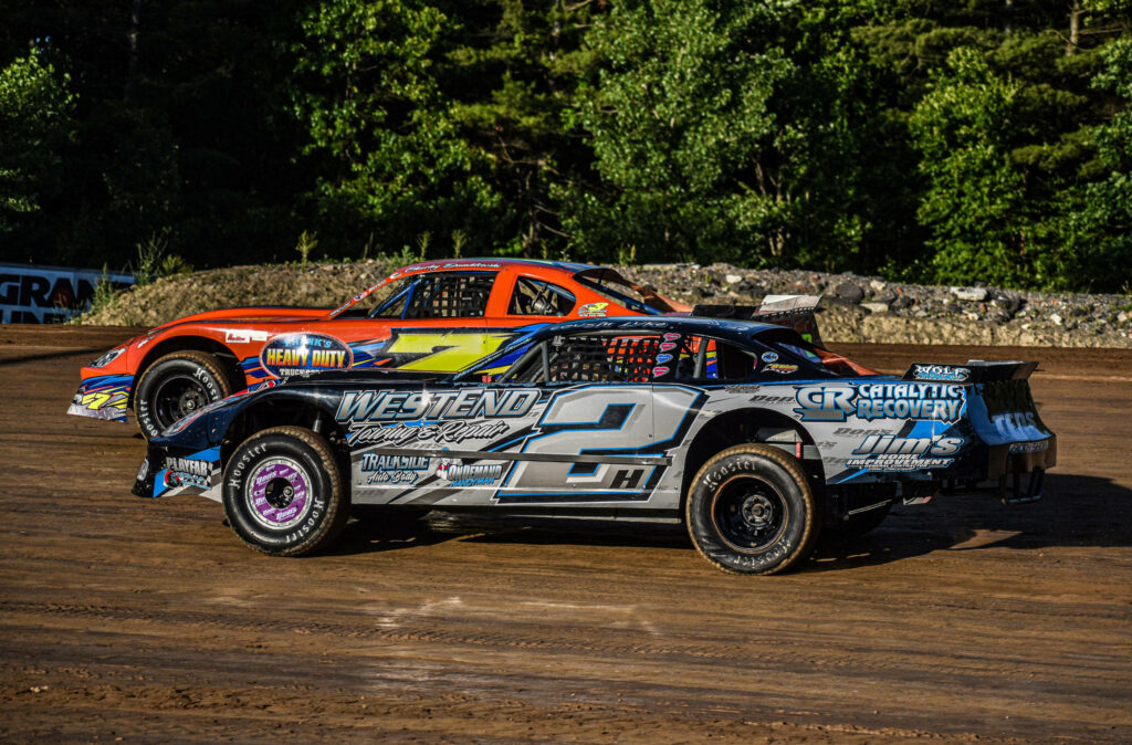 Two detailed dirt race cars speeding on sunlit dirt track with lush green trees background Wallpaper