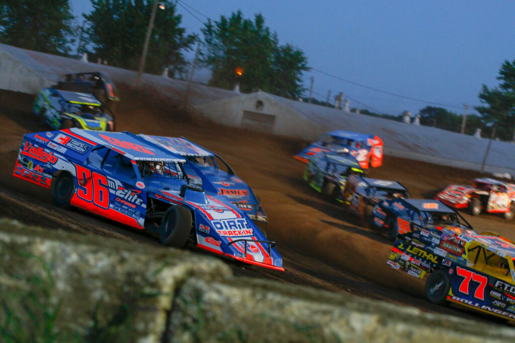 Vivid dirt cars race fast on muddy oval track at dusk under bright stadium lights intense action Wallpaper
