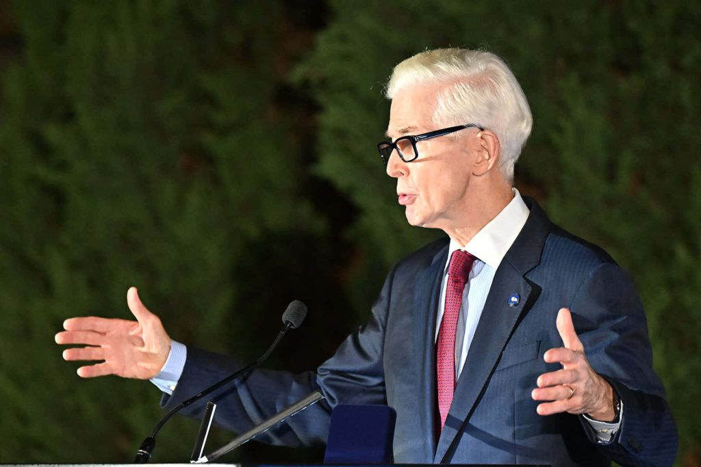 Distinguished man with silver hair glasses red tie speaking at podium with expressive gestures Wallpaper