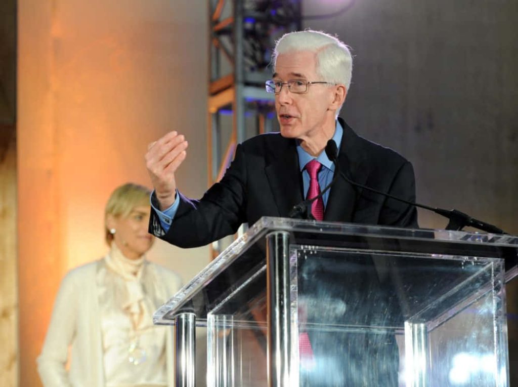 Distinguished speaker with white hair and glasses at acrylic podium in formal event setting Wallpaper