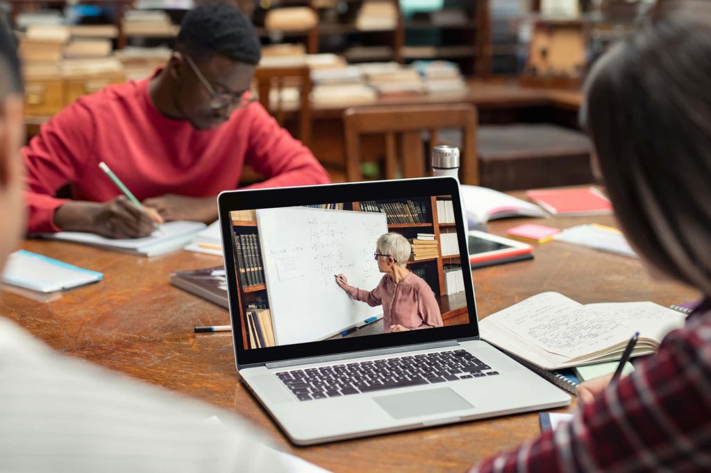 Diverse students studying at library table with laptop showing online math class instructor Wallpaper