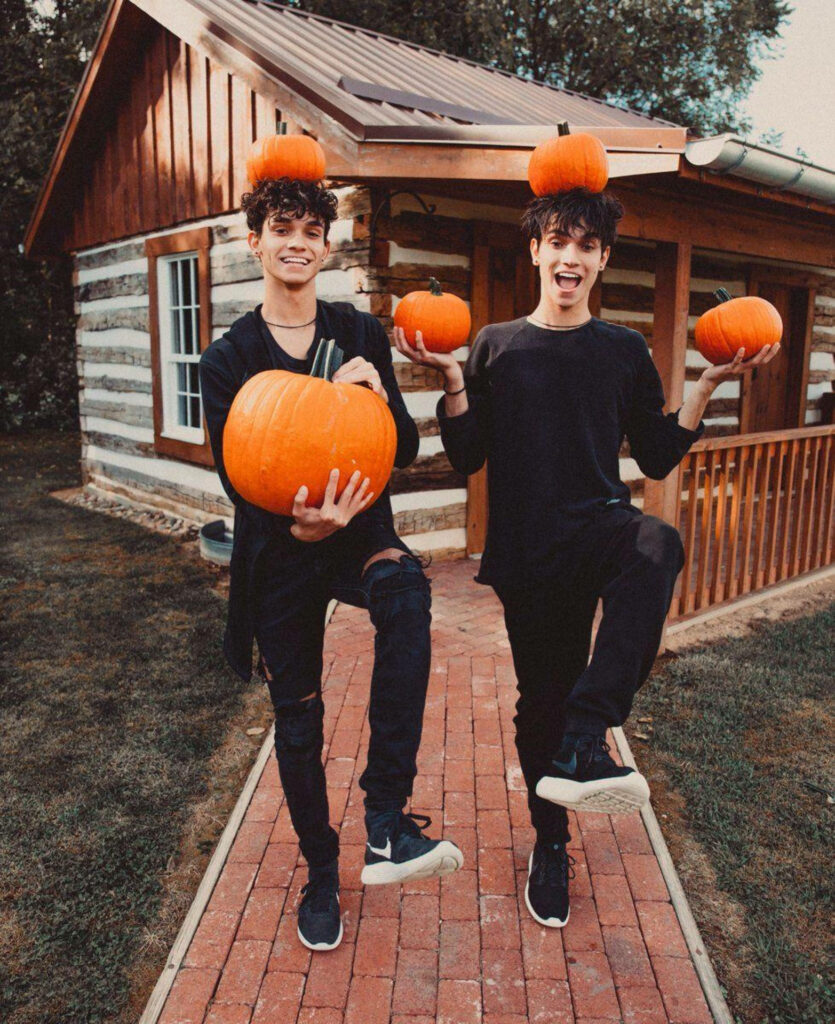 Dobre Brothers twins balancing pumpkins on heads outside rustic cabin autumn Halloween vibes wallpaper