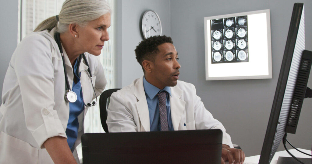 Two doctors in white coats reviewing medical data on computer with brain scans in clinical office Wallpaper