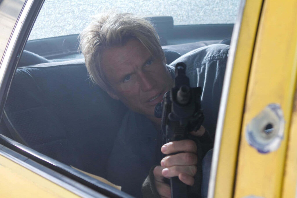 Dolph Lundgren aims rifle crouched in car backseat intense action scene blue yellow tones wallpaper