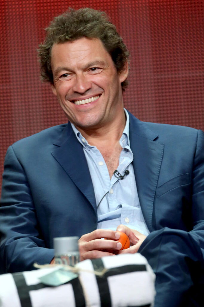 Dominic West in smart blue suit smiling warmly against textured red background wallpaper