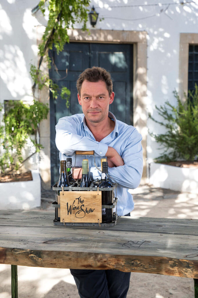 Dominic West outdoors leaning on wooden table with Wine Show bottles and rustic white building background Wallpaper
