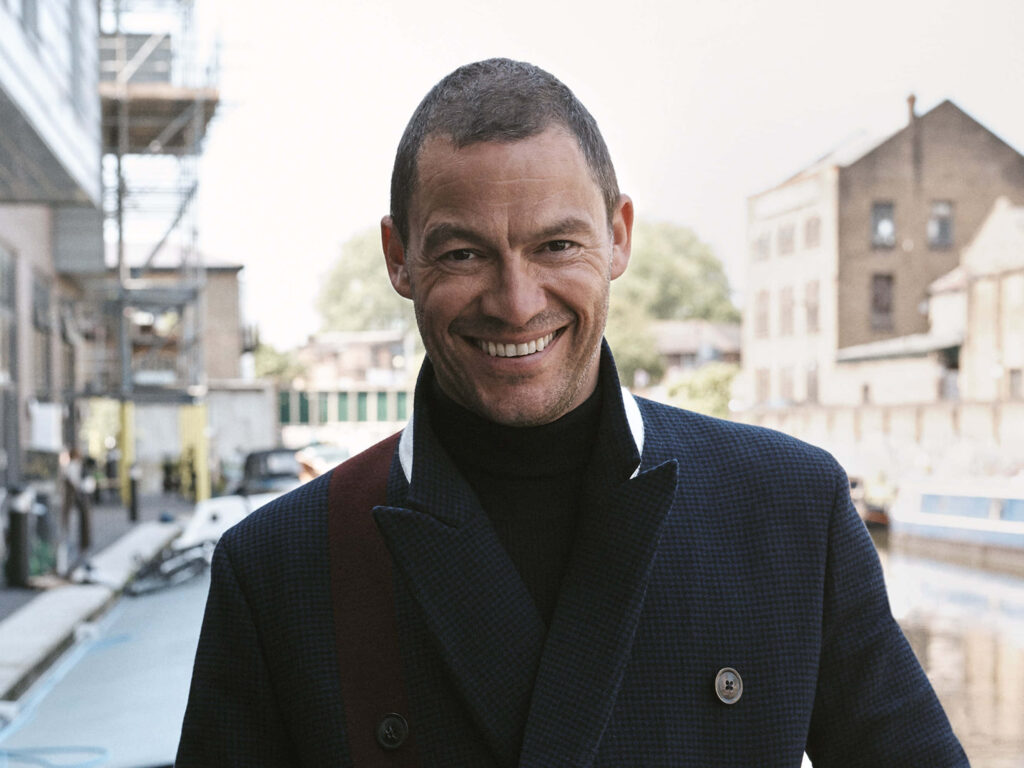 Dominic West in dark checkered coat by serene urban riverside with confident smile and soft light Wallpaper