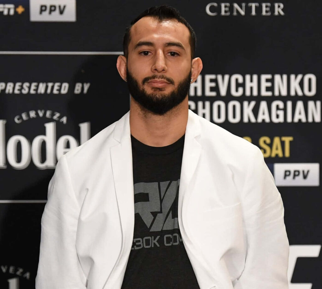 Dominick Reyes in white blazer black Reebok shirt at MMA event with sponsor backdrop and lighting Wallpaper