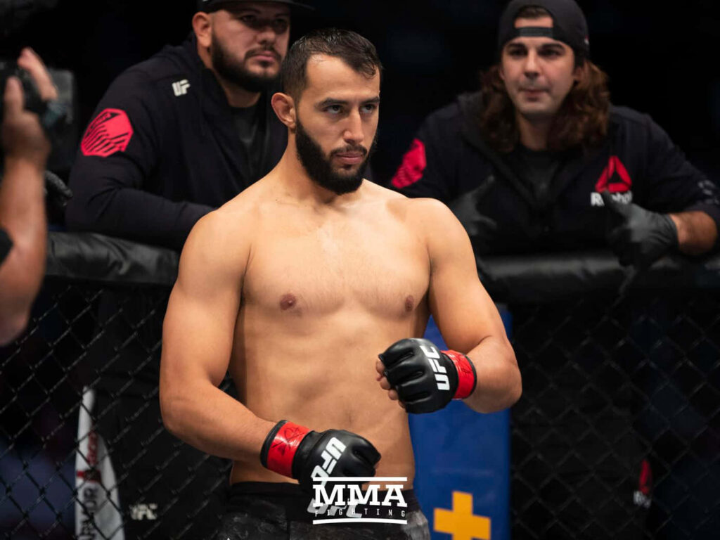 Dominick Reyes focused in MMA cage with athletic stance under lights and team watching intensely Wallpaper