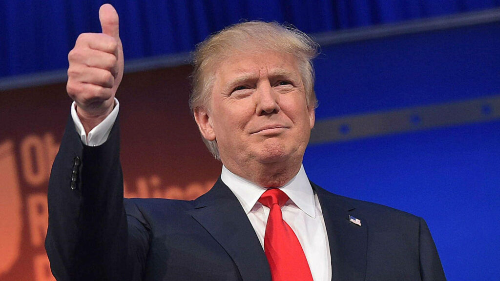 Donald Trump in sharp suit and red tie giving thumbs up with confident smirk on blue background Wallpaper