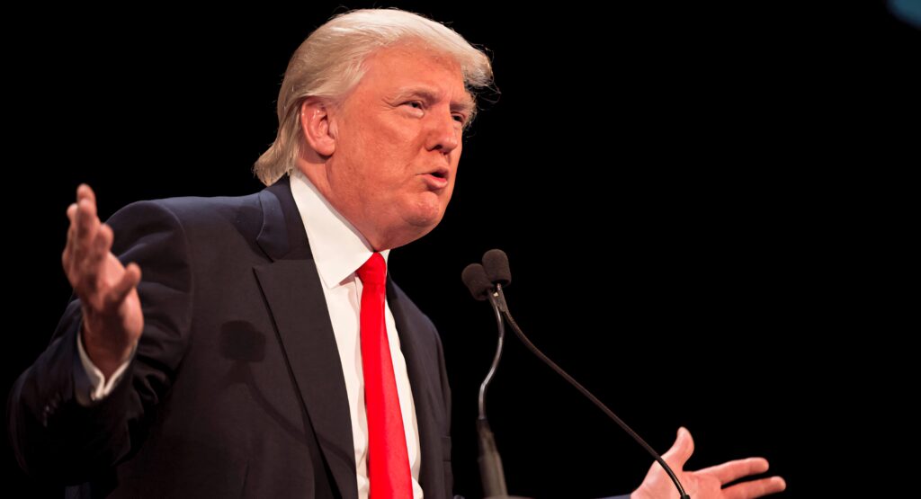 Donald Trump speaking at podium with expressive gestures dark background dramatic lighting wallpaper