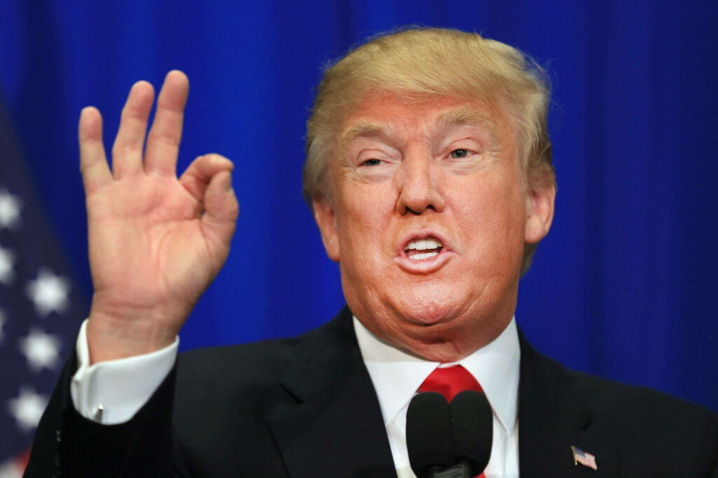 Donald Trump speaking with iconic hand gesture in suit against blue backdrop and American flag Wallpaper