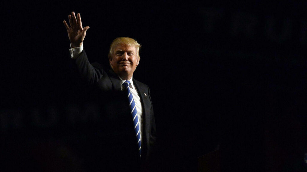 Donald Trump raising hand against dark background with dramatic lighting highlighting suit and hair Wallpaper