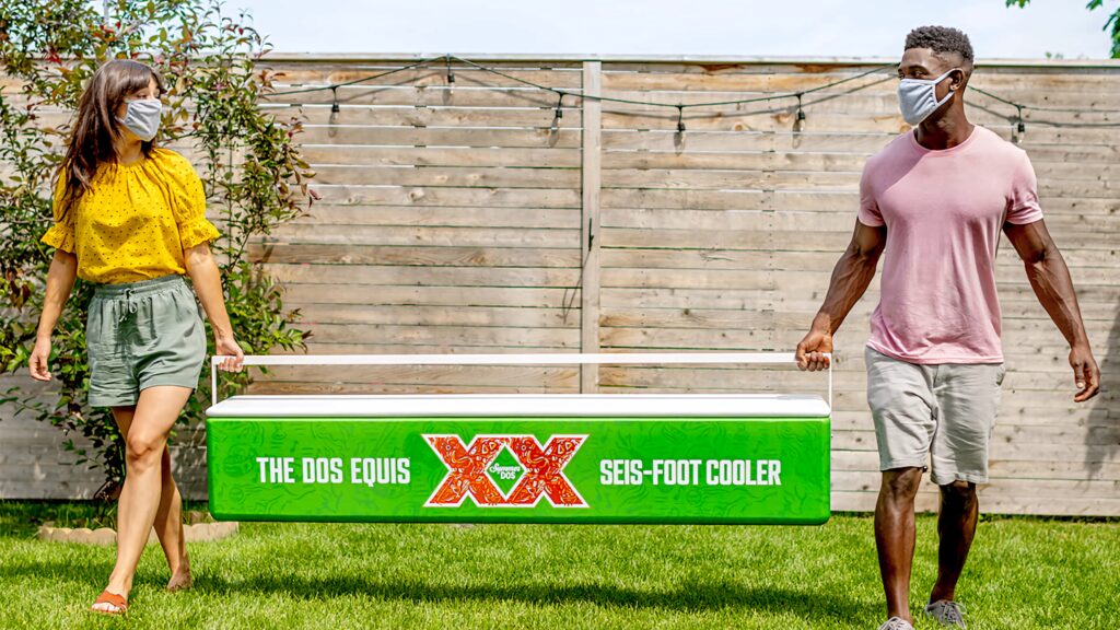 Two people in summer masks carrying green Dos Equis Seis cooler in sunny backyard background Wallpaper