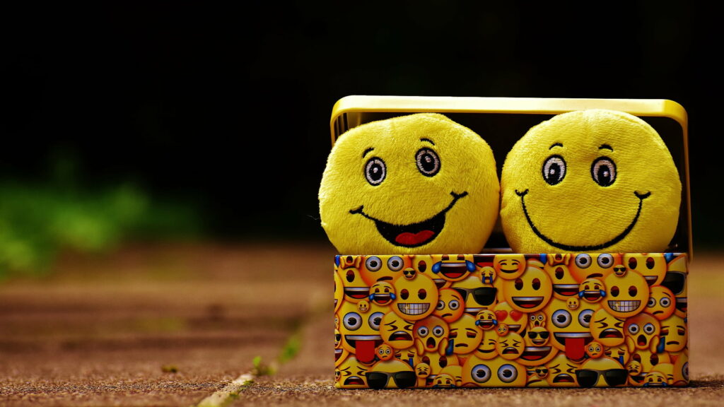 Two fuzzy yellow smiley plush balls peeking from emoji-decorated box vibrant wallpaper background