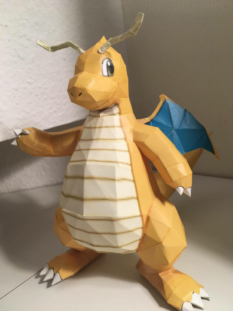 Papercraft Dragonite model with orange cream body and blue wings on neutral indoor wallpaper background