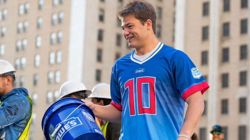 Drake Maye smiling in blue number 10 jersey with vibrant urban background and lively atmosphere Wallpaper