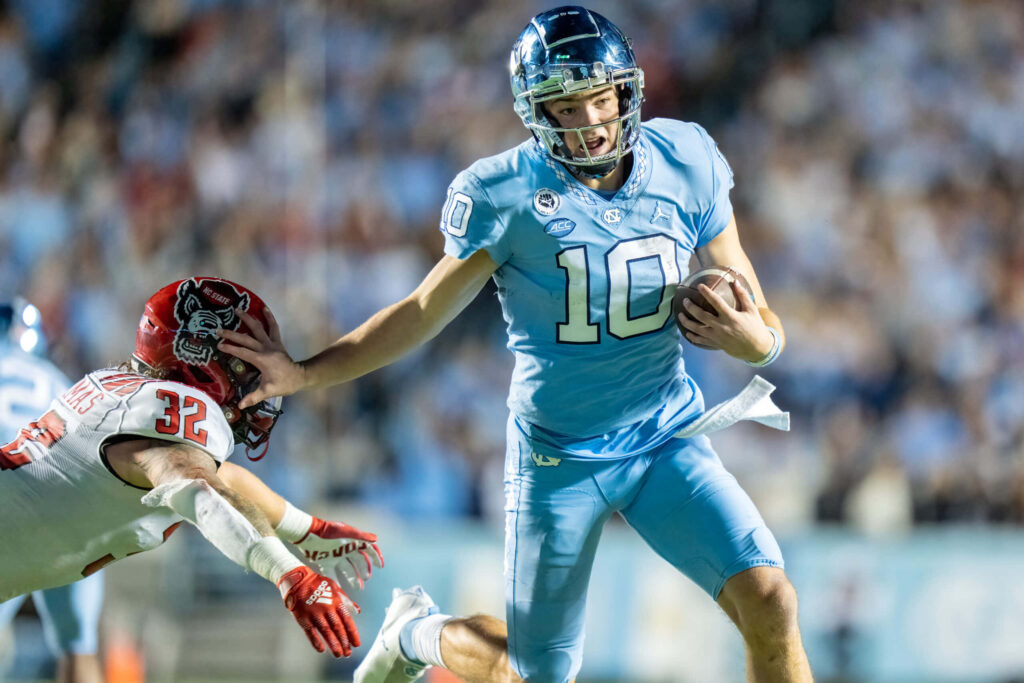 Drake Maye in light blue uniform dodging tackle showing agility and determination football action Wallpaper