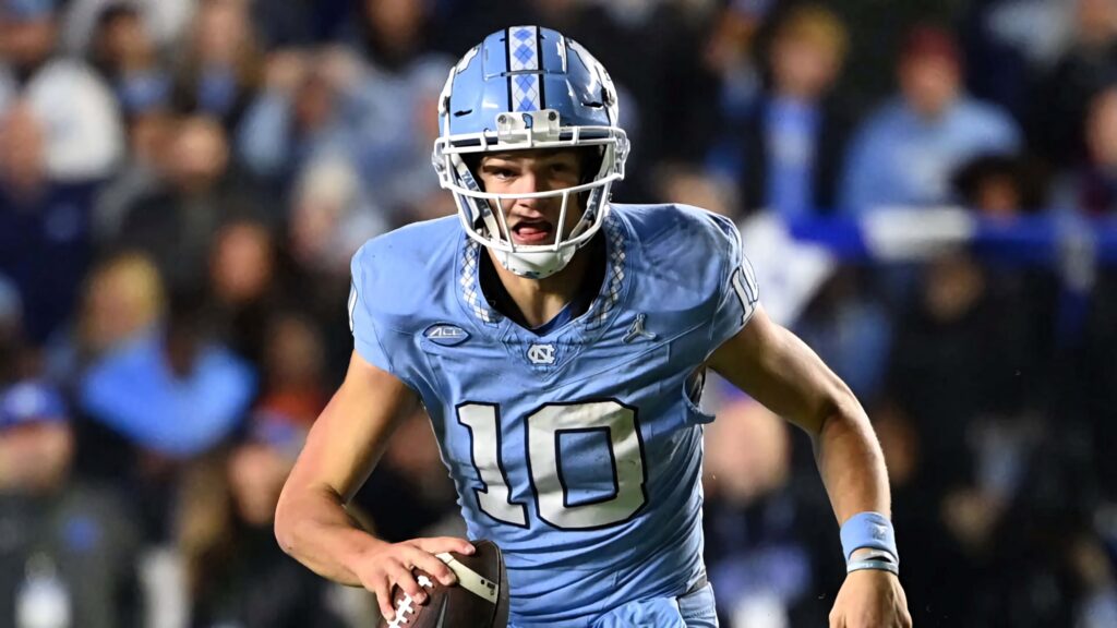 Quarterback Drake Maye in light blue North Carolina jersey throws pass under bright stadium lights Wallpaper