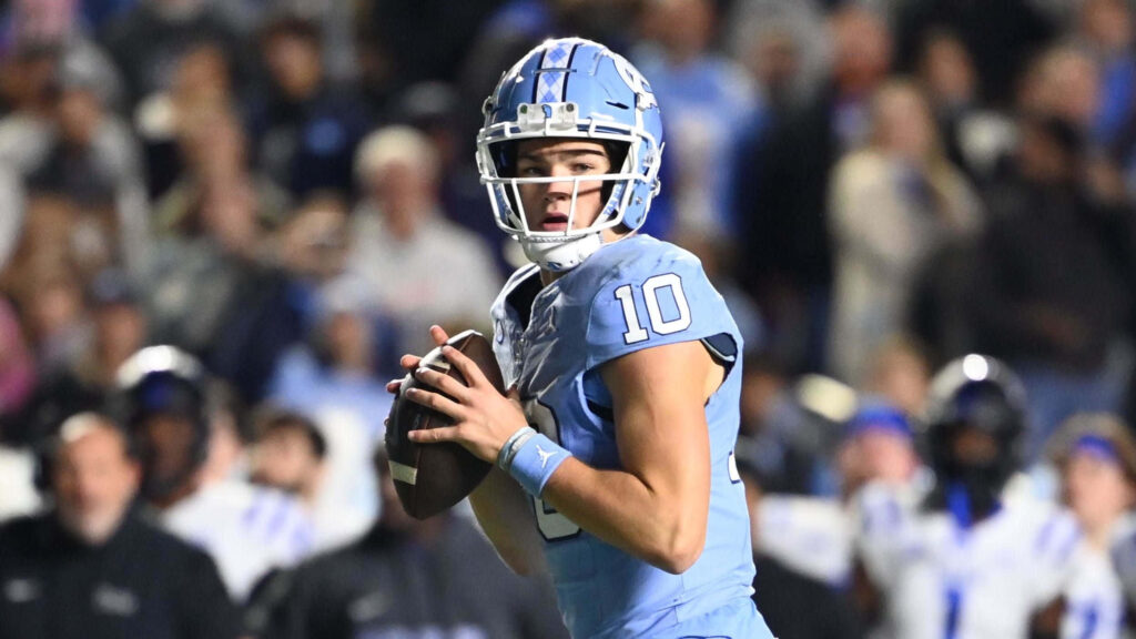 Drake Maye in light blue North Carolina jersey poised to throw football in blurred stadium background Wallpaper