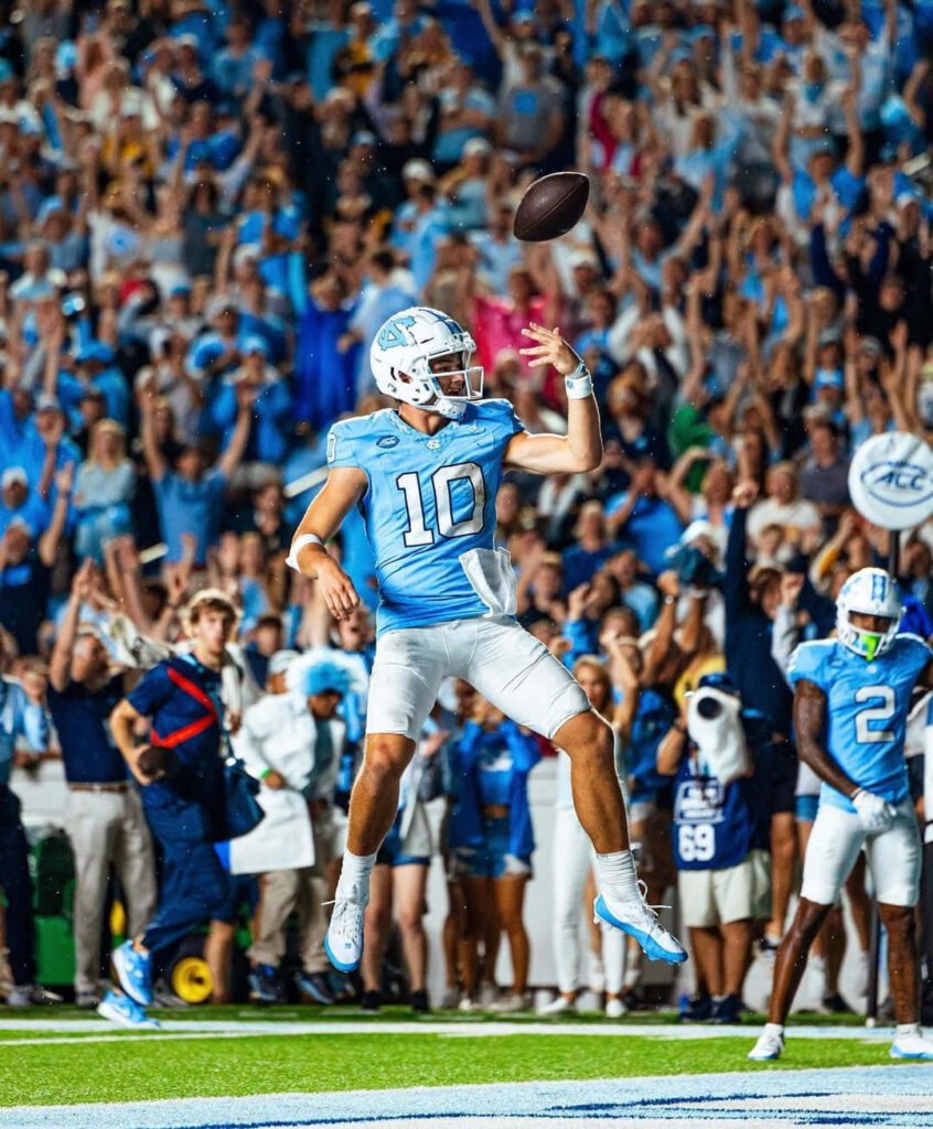 Drake Maye in number 10 jersey throwing football mid-air with packed stadium and fans background Wallpaper
