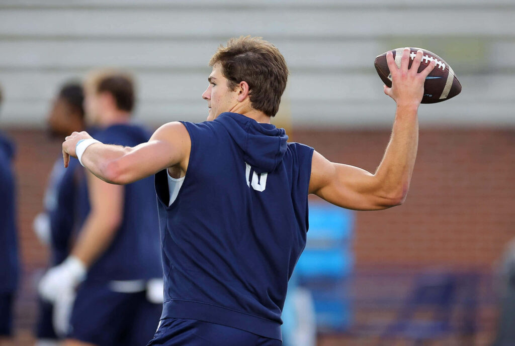 Drake Maye in navy hoodie focused throwing football during intense pass practice on training field Wallpaper