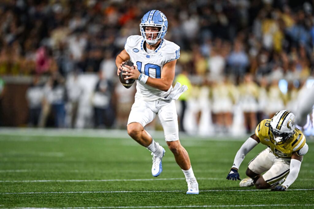 Drake Maye in white blue uniform evading tackle under stadium lights during intense football game Wallpaper
