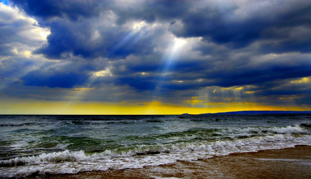 Dramatic 4K coastal wallpaper with sunbeams piercing storm clouds over tranquil ocean waves