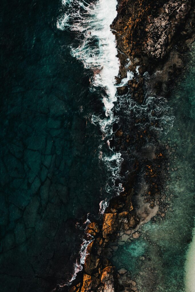 Aerial view of rugged coastline with deep blue sea crashing against textured rocks wallpaper background
