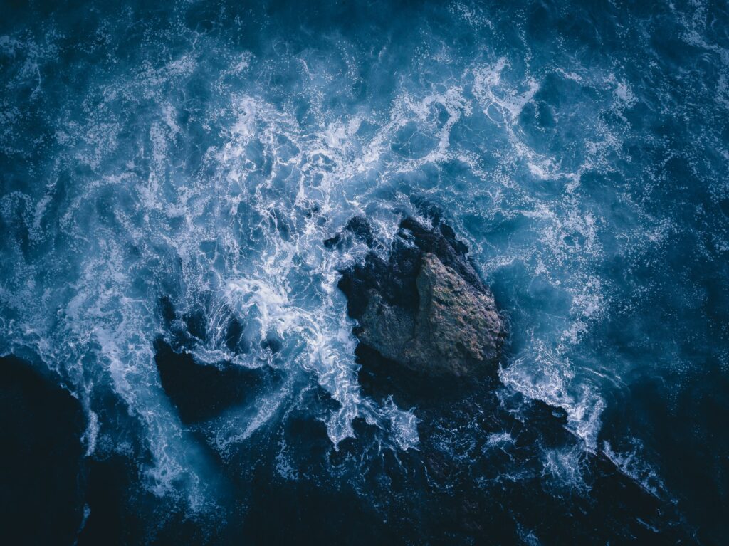 Aerial view of ocean waves crashing on rugged stone with white foam in deep blue water wallpaper