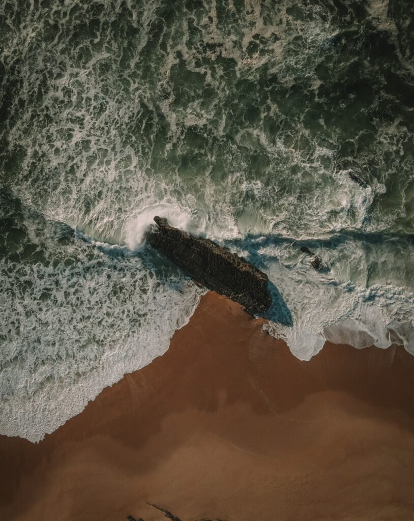 Aerial view of powerful sea waves crashing on sandy beach around dark rocky outcrop wallpaper background