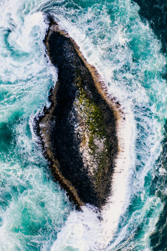 Aerial view of rugged rock island surrounded by swirling turquoise sea and white foam wallpaper
