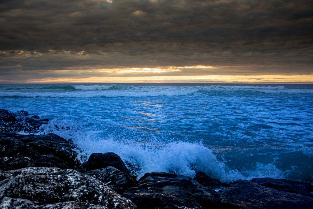 Dramatic blue sea waves crashing on dark rugged rocks with white spray under sunset sky Wallpaper
