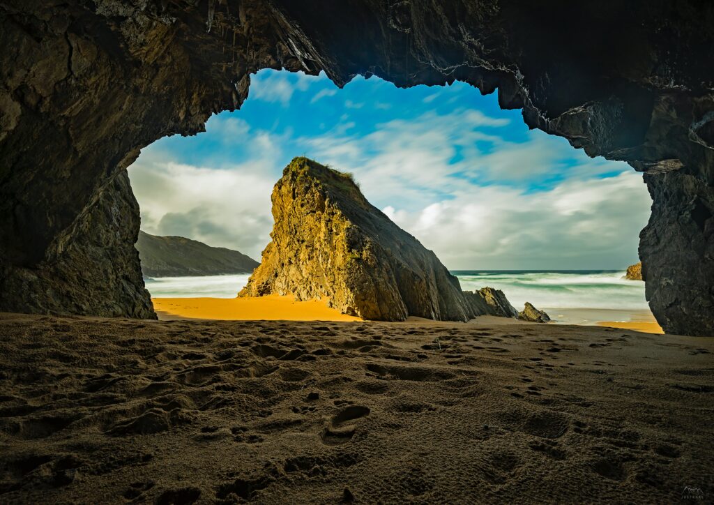 View from dark cave to sunlit beach with towering rock formation and golden sand wallpaper background