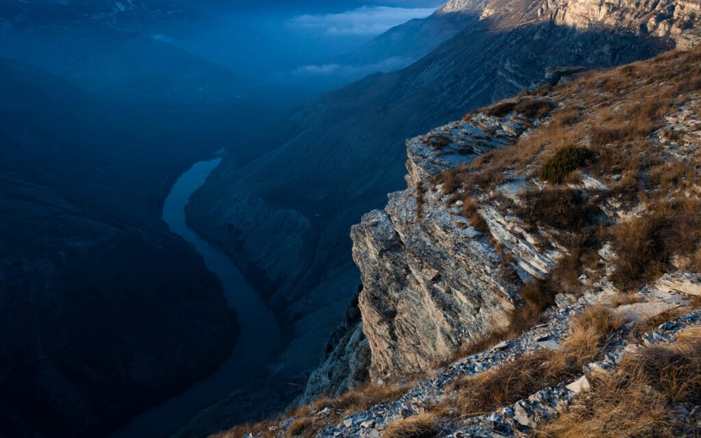 Aerial view of rugged cliff with sun-faded grass overlooking winding river and misty mountains Wallpaper