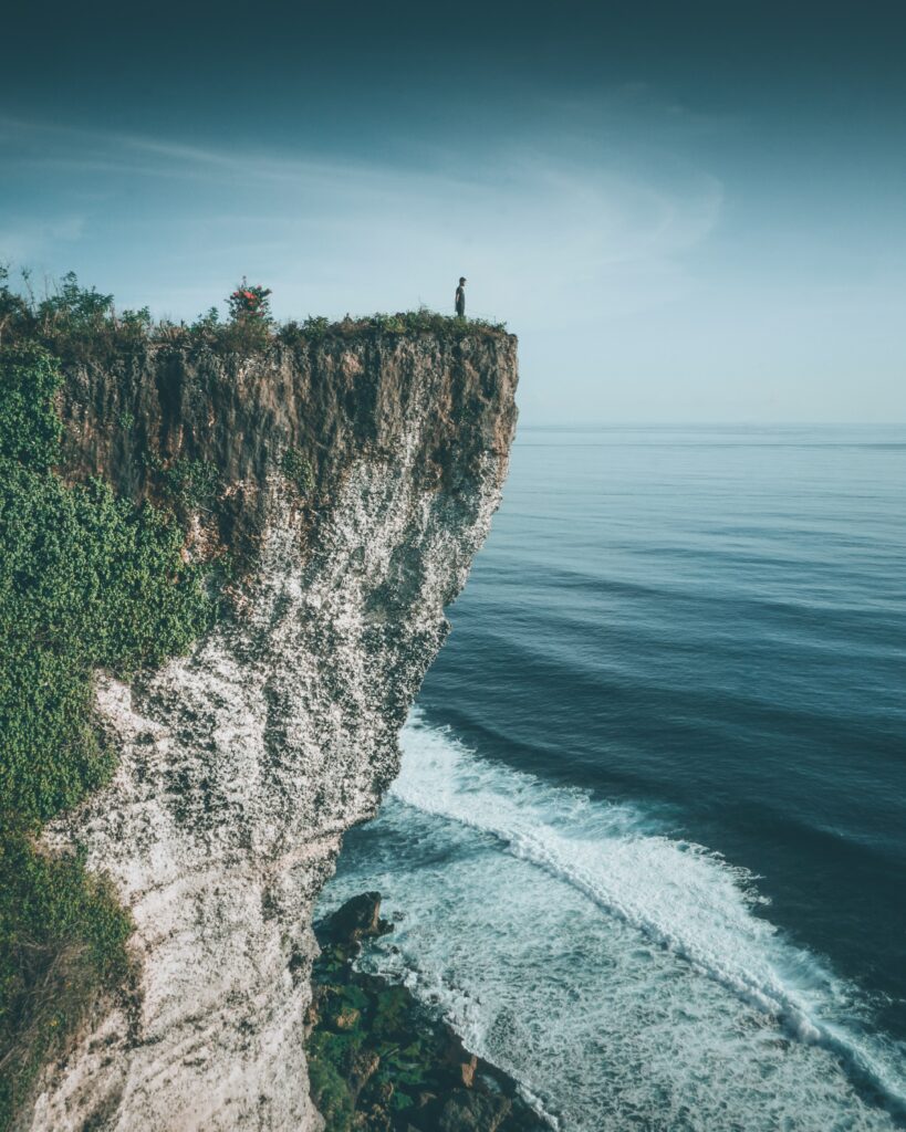 Dramatic cliff with lush greenery and lone silhouette overlooking serene blue sea waves wallpaper