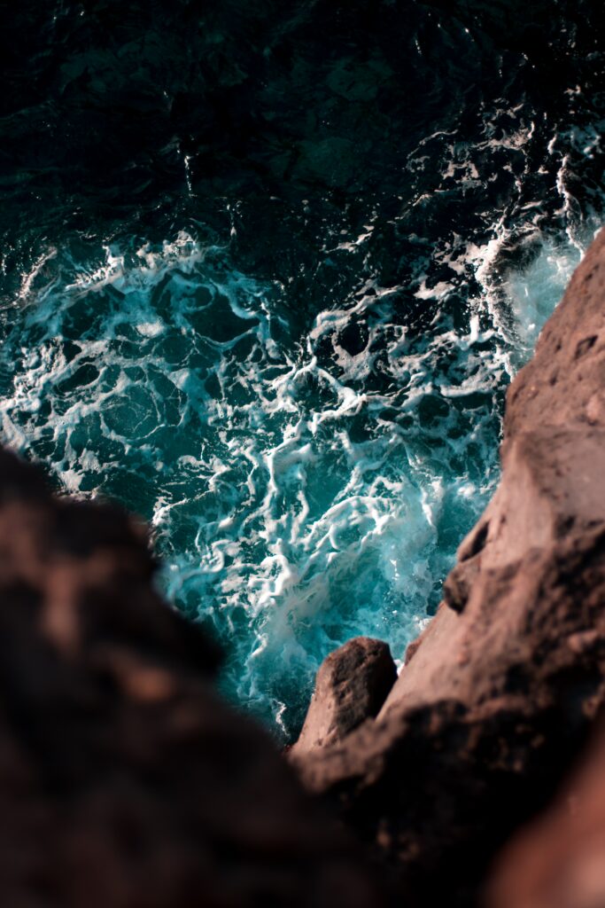 Cliffside view of foamy waves crashing on rugged rocks with deep blue sea and warm stone tones Wallpaper