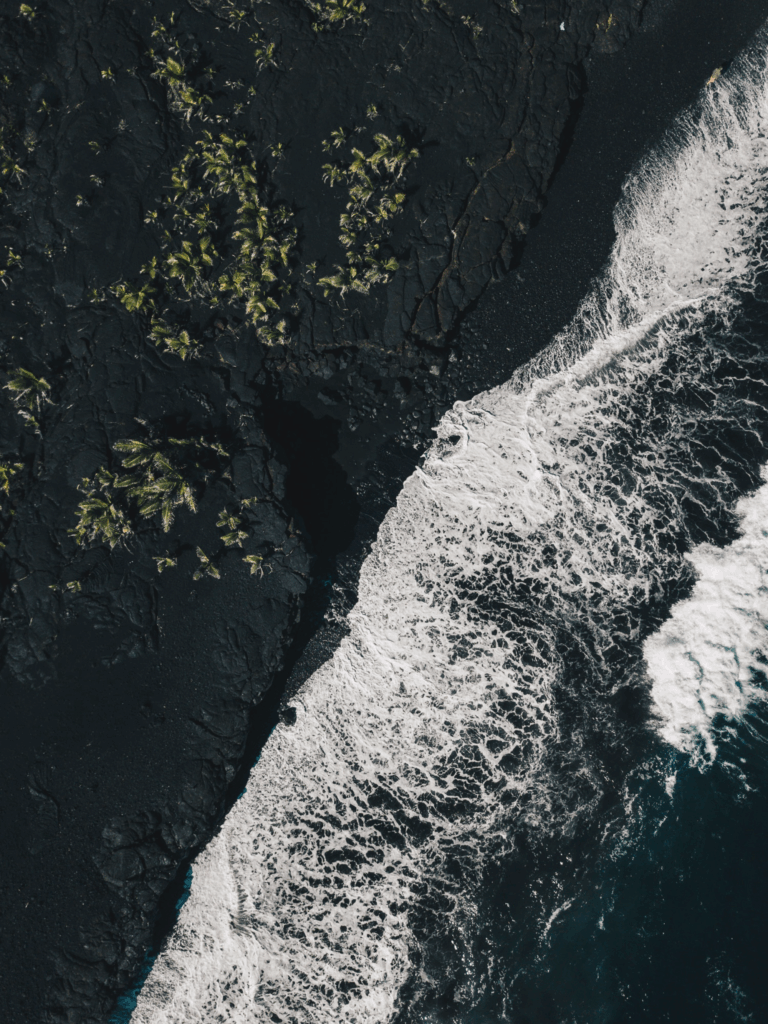 Aerial view of foamy ocean waves crashing on rugged volcanic rocks with lush green coastline Wallpaper